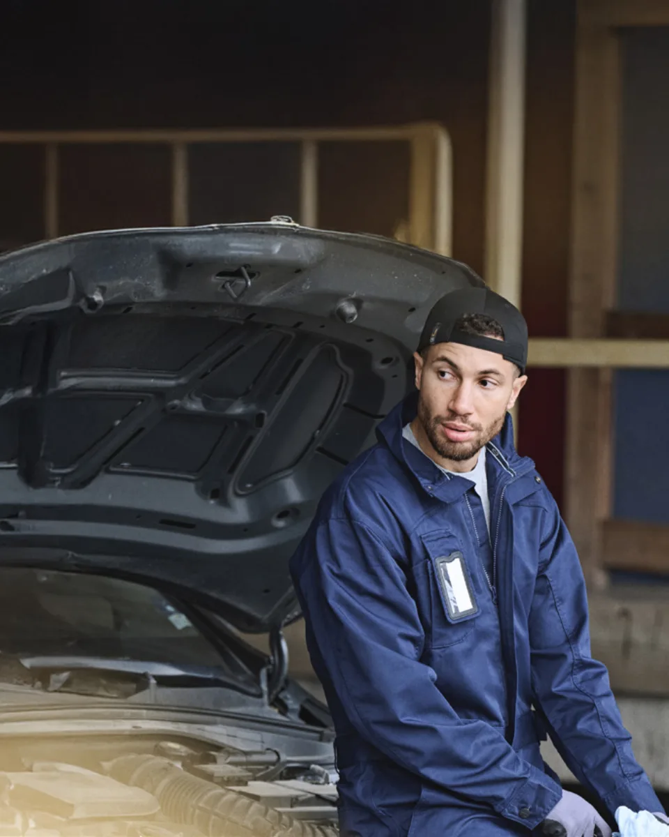 Mekaniker i marinblå arbetsjacka och keps sitter vid en bil med öppen motorhuv