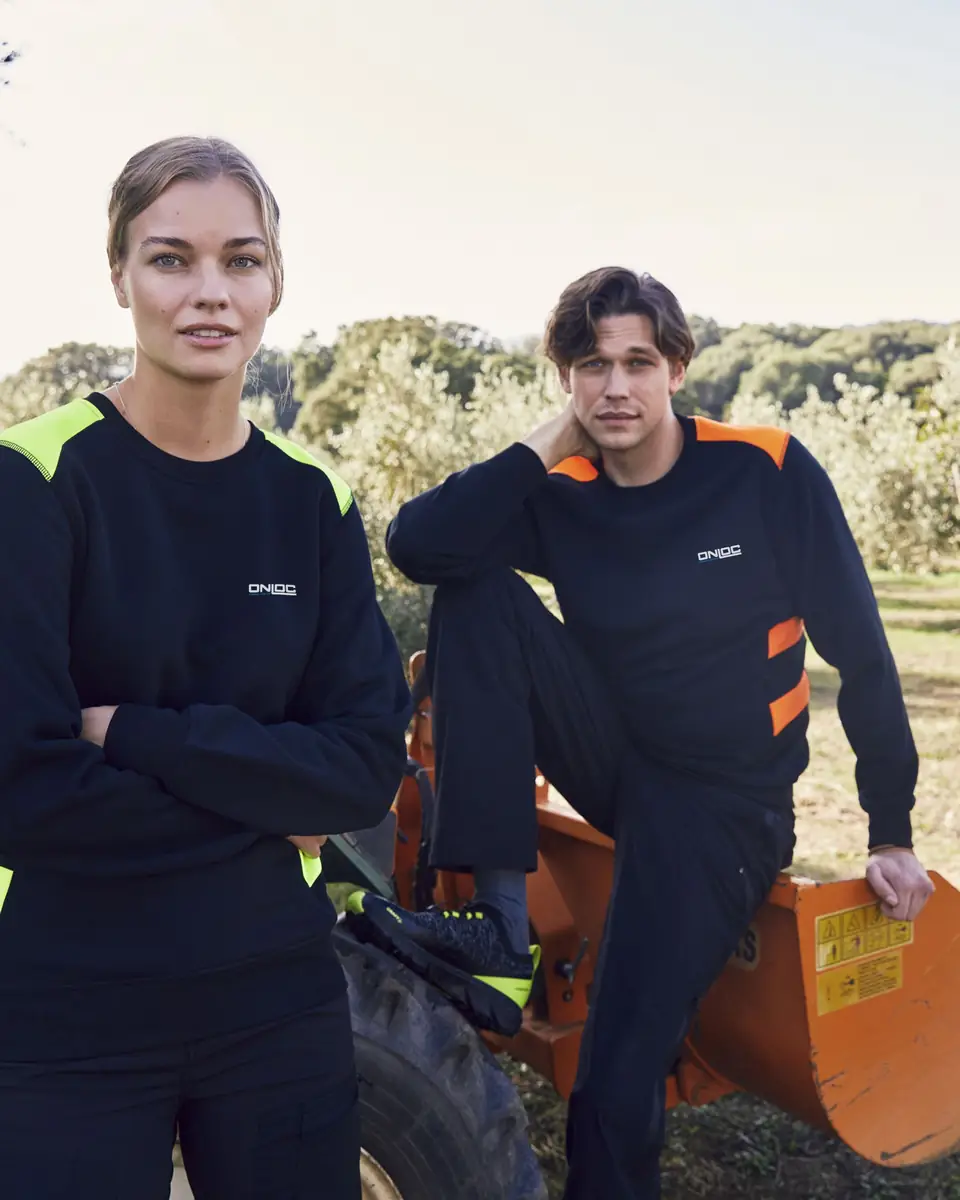 Två lagerarbetare i svarta tröjor med gula och orange kontrastpartier och logotyp på bröstet